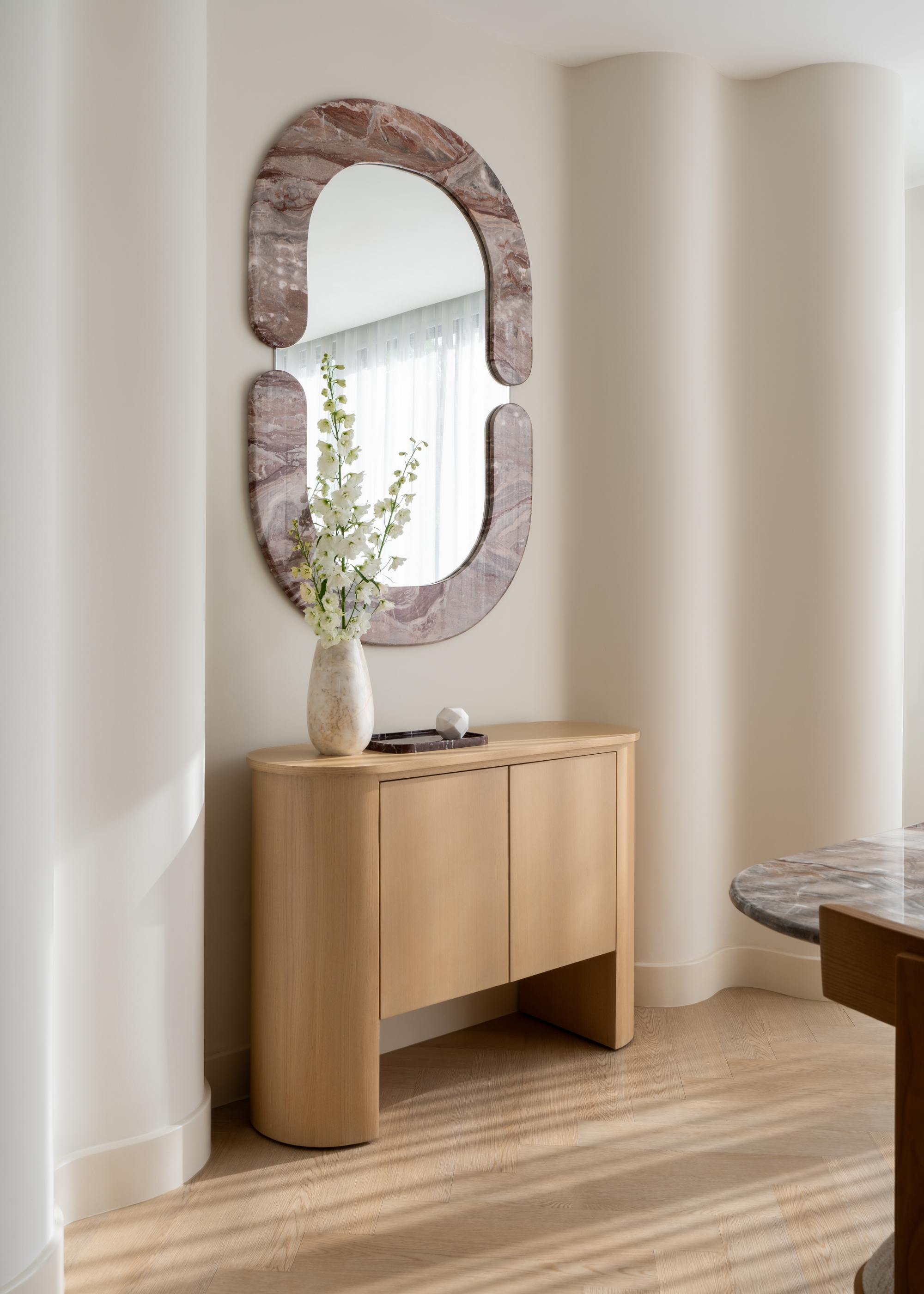 A living room area with a wood sideboard, a marble framed wall mirror, and a vase of white flowers