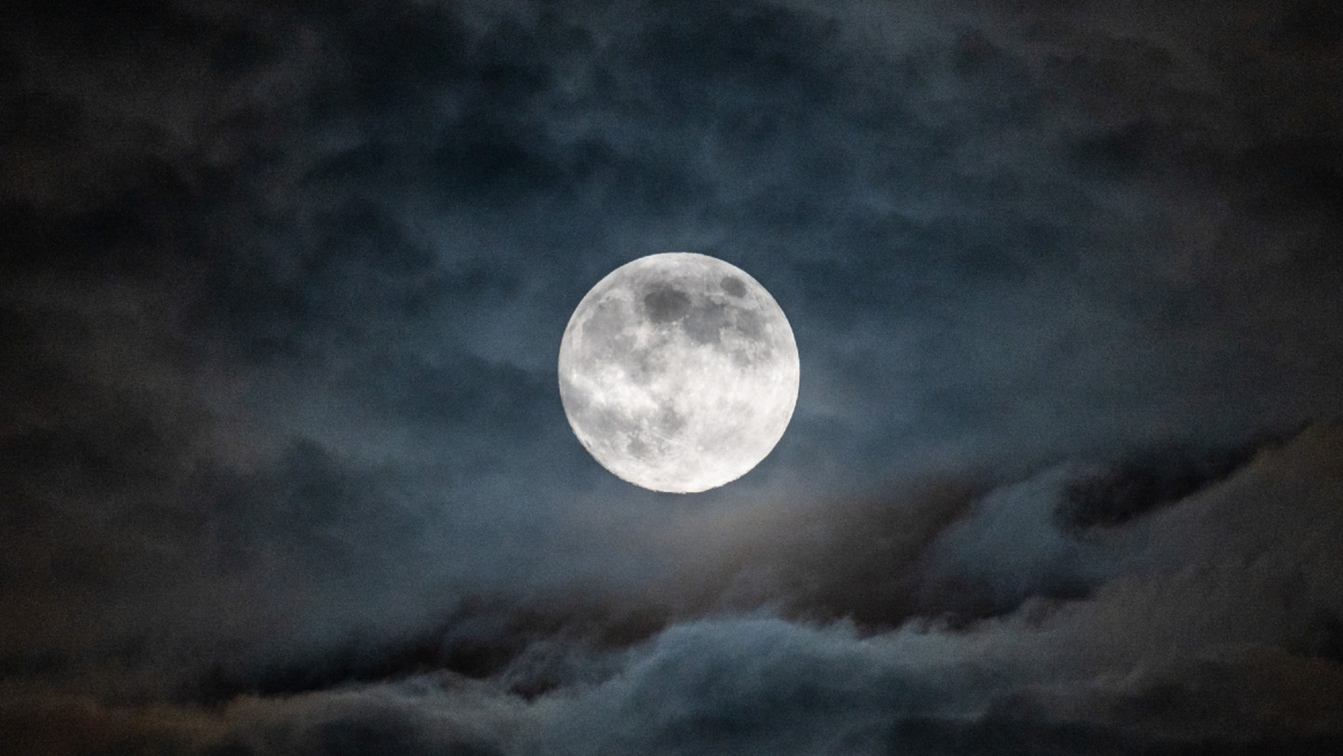 A full moon is pictured in a cloudy night sky, with the ancient lava plains known as lunar maria darkening its otherwise pearlescent surface.