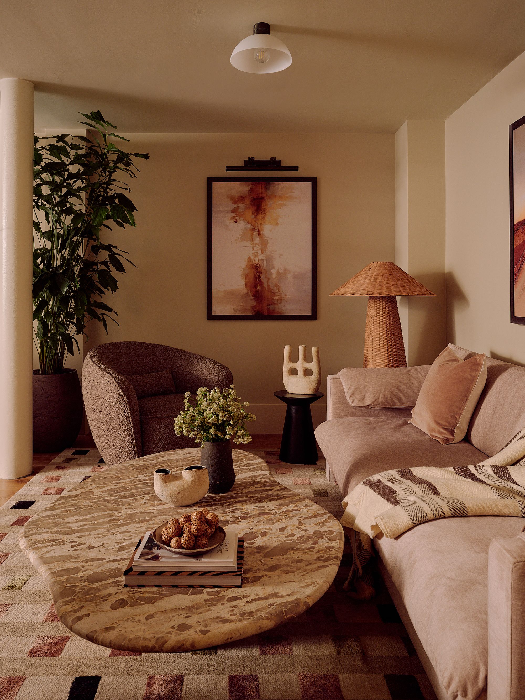 neutral living room with soft brown linen sofa with throw and cushion, rattan floor lamp, side table with sculpture, brown tub accent chair, stone organic-shaped coffee table styled with vases and books, checkered rug, large potted tree in the corner and artwork on the wall