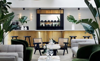 Bar area in the MyChelsea hotel. Golden bar, with a white marble countertop, and a black and golden shelf behind it with a mirror. Sitting is in gray and olive green upholstered chairs, in combination with black and gold wooden chairs. White marble coffee tables are in between.