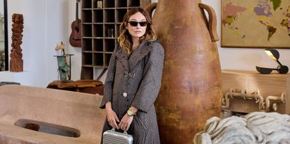 A young woman dressed in a textured black coat with white detailing and a silver brooch and tortoise sunglasses stands with a grey bag in her hands in a furniture showroom filled with vintage items.