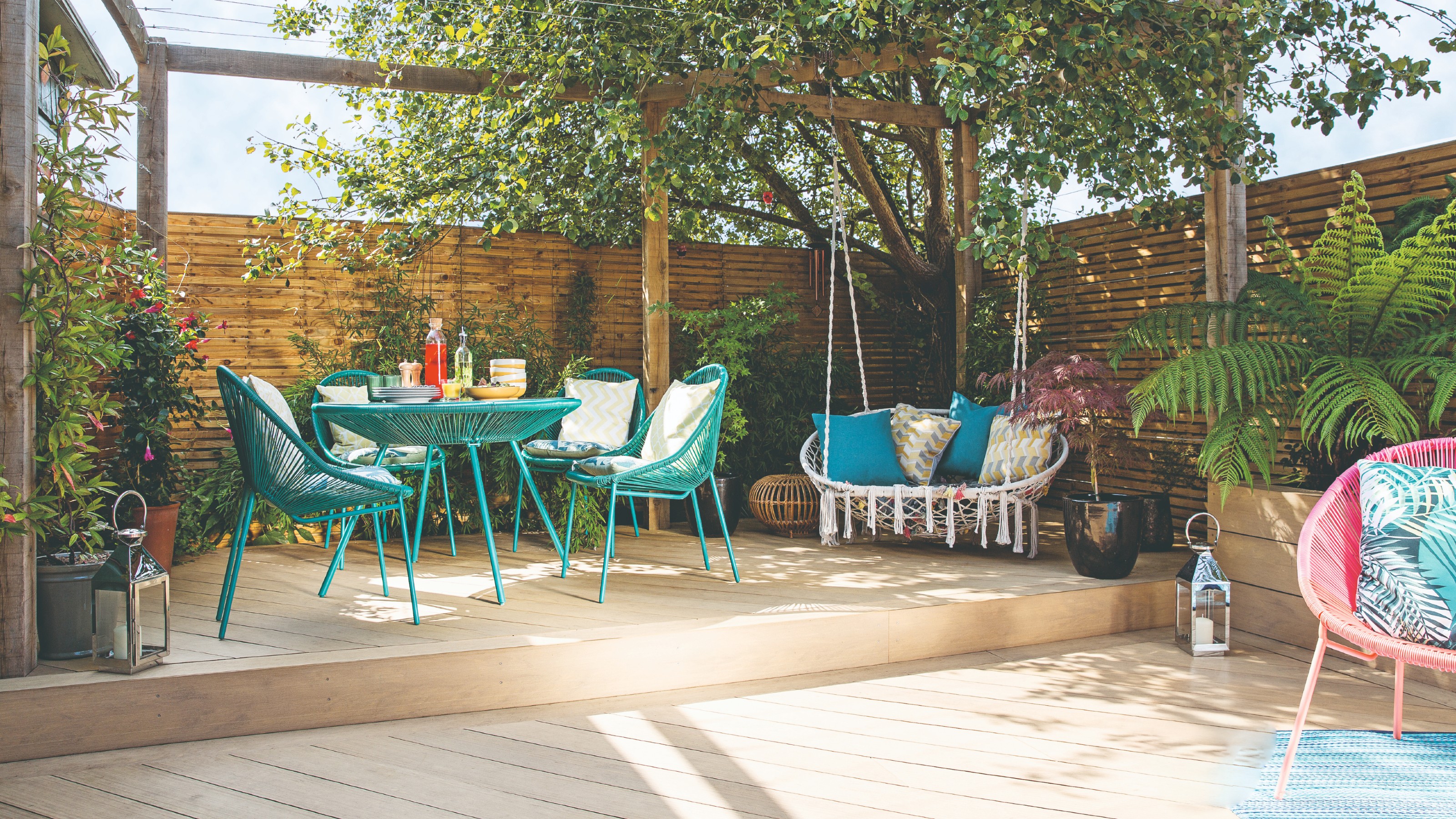 A garden with an elevated platform with a turquoise dining set and a hanging chair and a pink Acapulco-style lounge chair on the lower end of the garden