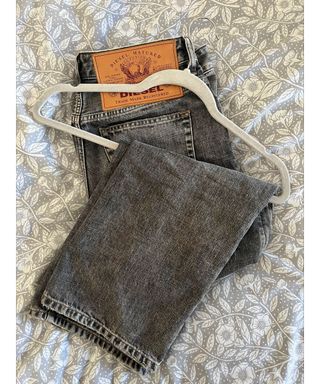 A pair of dark grey Diesel jeans on a grey velvet hanger on grey flowery bedding