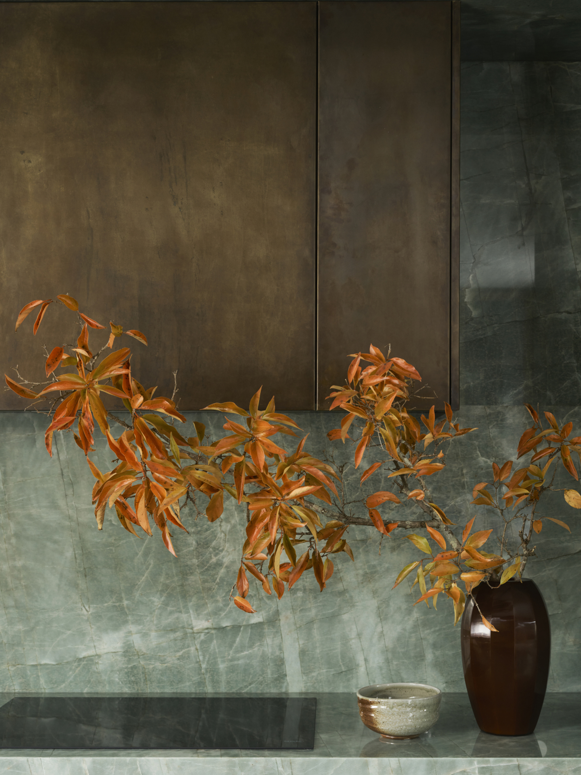 Close-up image of a kitchen counter and stove top with aged brass cabinets above it. The countertop and backsplash are made out of a teal stone material. There is a vase of bright orange flowers.