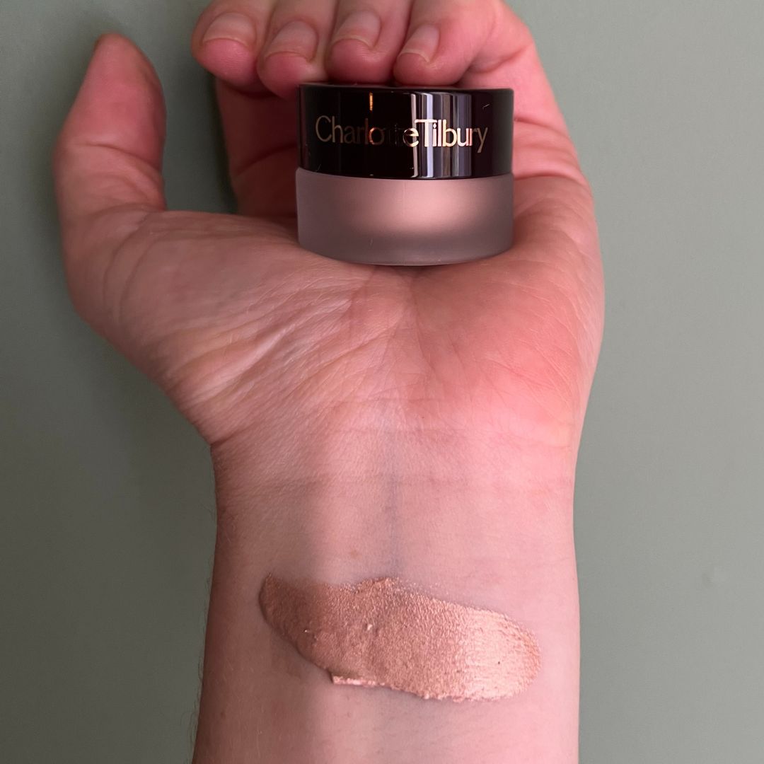 a hand on a green background holding charlotte tilbury eyes to mesmerise cream eyeshadow with a swatch of the eyeshadow on the wrist 