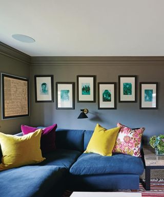 A living area with black and white framed wall prints on the dark gray wall, a navy blue couch with yellow and purple throw pillows, and a dark gray wooden side table, and a large white ceiling