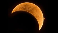 a close up view of the sun during a partial solar eclipse with the new moon in the lower left corner obscuring a large portion of the sun. 