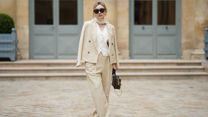 A guest wears black sunglasses, gold earrings, a white silk scarf, a gold chain pendant necklace, a white tweed buttoned V-neck gilet jacket, a beige linen buttoned blazer jacket, matching beige large suit pants, a brown shiny LV monogram print pattern in coated canvas zipper Petite Malle handbag from Louis Vuitton, black shiny leather platform soles / block heels ankle boots , outside the COS show, on April 26, 2023 in Paris, France.
