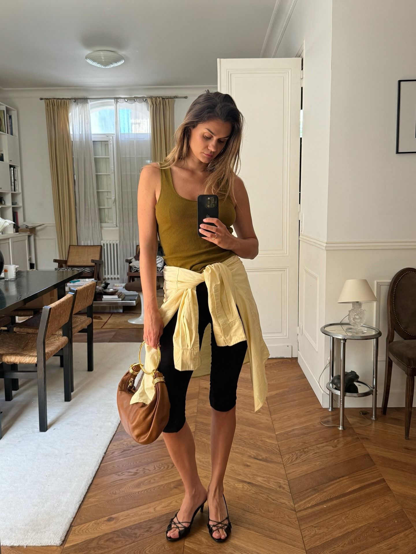 Influencer @annelauremais takes a mirror selfie wearing a khaki tank top, black capris and heeled sandals. She wears a yellow shirt looped around her waist and holds a Chloe bracelet bag.