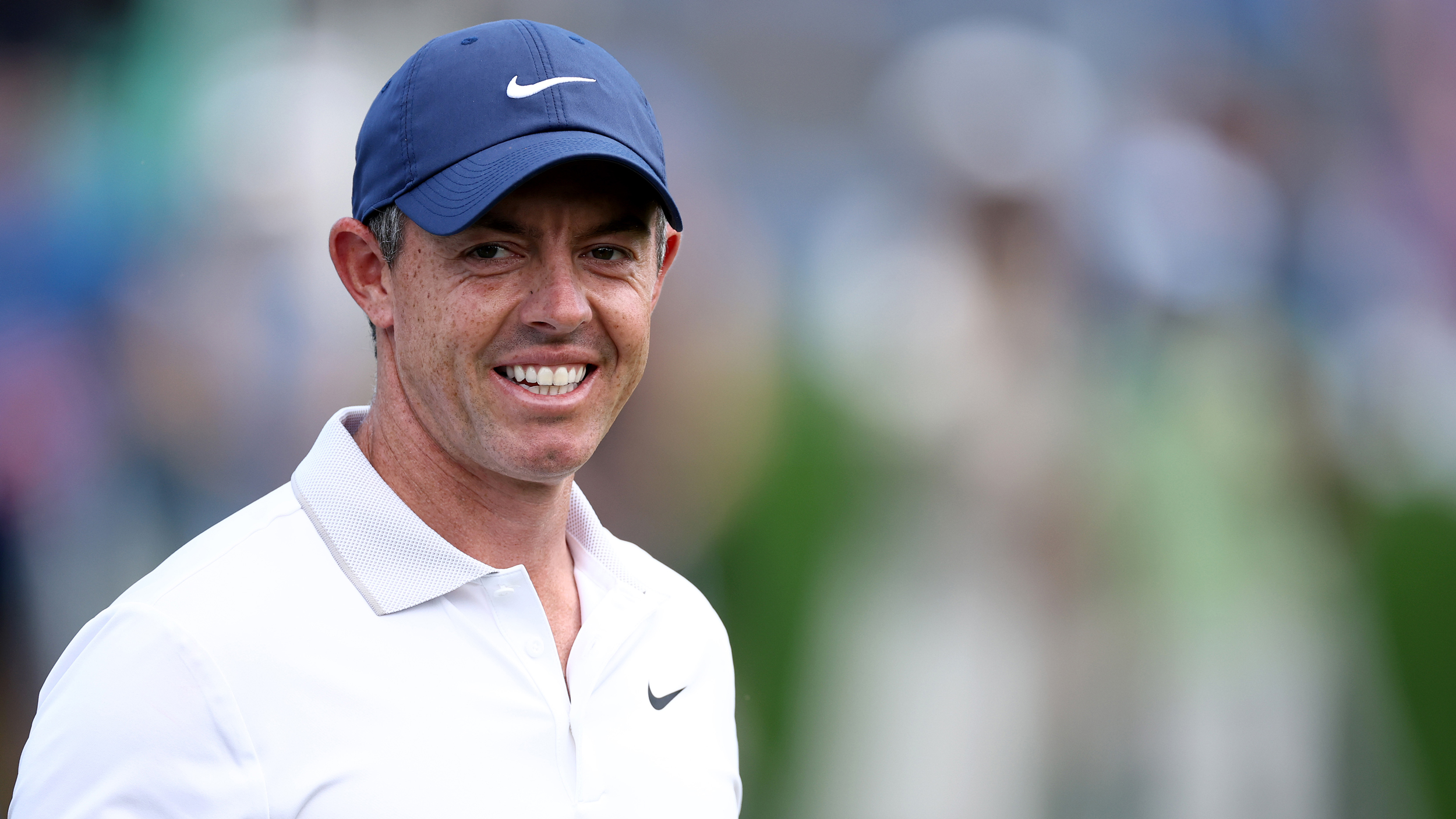 Rory McIlroy smiles while wearing a white polo and navy blue cap