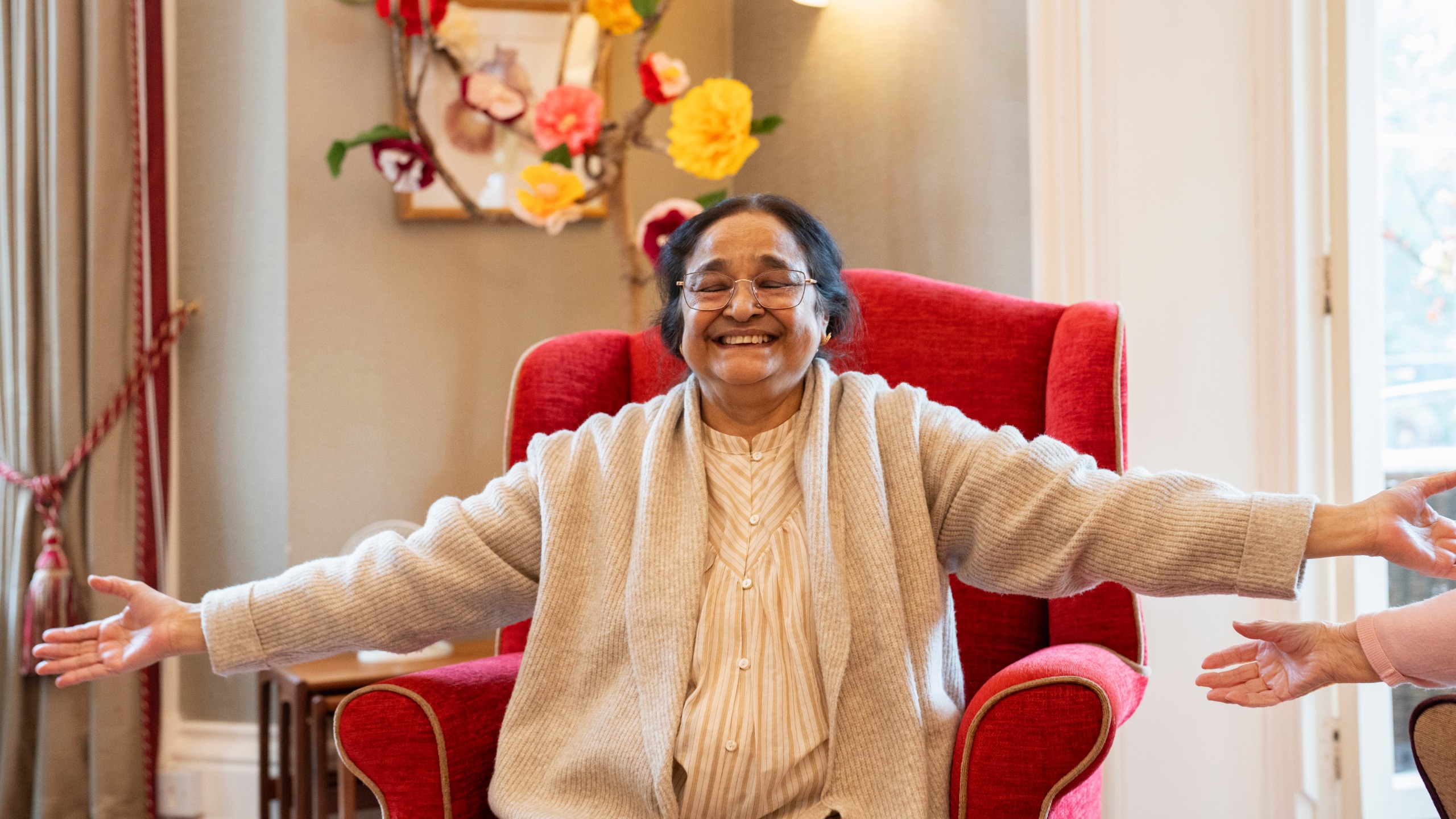 A senior woman sits in a comfy armchair smiling and holding her arms out to the sides at shoulder height.
