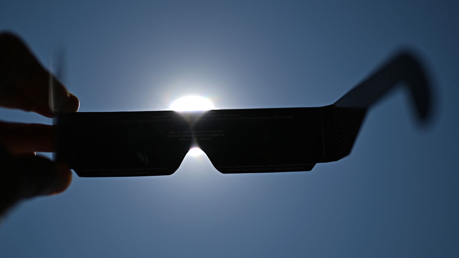 A pair of eclipse glasses are held up close to the sun, which shines in a blue sky.
