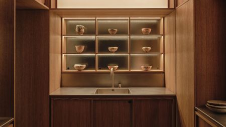 A Japanese kitchen with a steel sink on a stone slab, wood cabinets, and a display of ceramics in backlit compartments