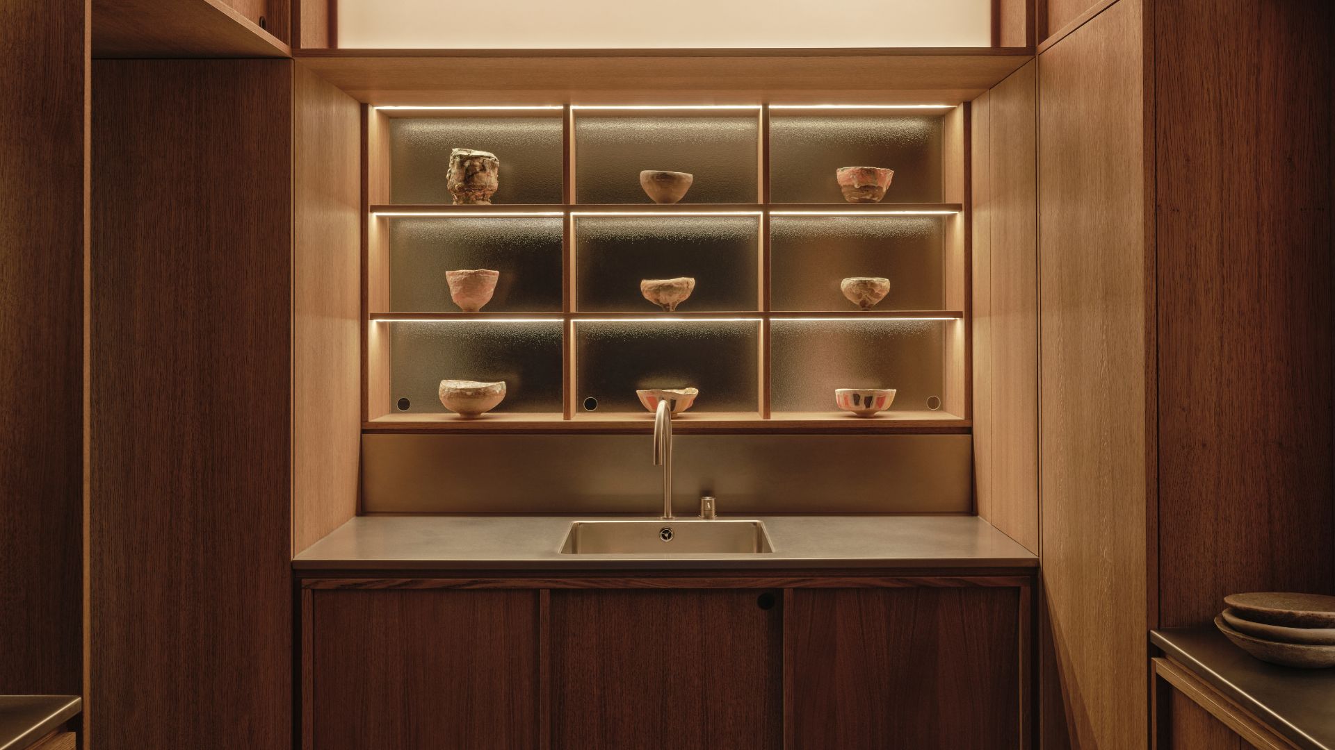 A Japanese kitchen with a steel sink on a stone slab, wood cabinets, and a display of ceramics in backlit compartments
