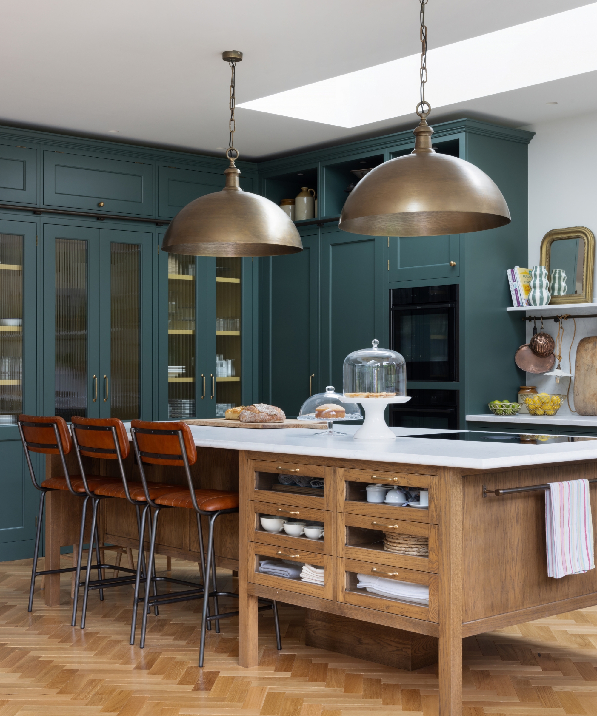 Dark green shaker kitchen with brass dome pendants, a wooden island with open shelving, and leather bar stools.