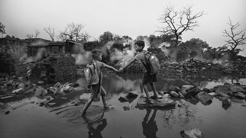 Two boys with backpacks hold hands while crossing a rocky, flooded area. Bare trees and ruins in the background create a somber and resilient mood