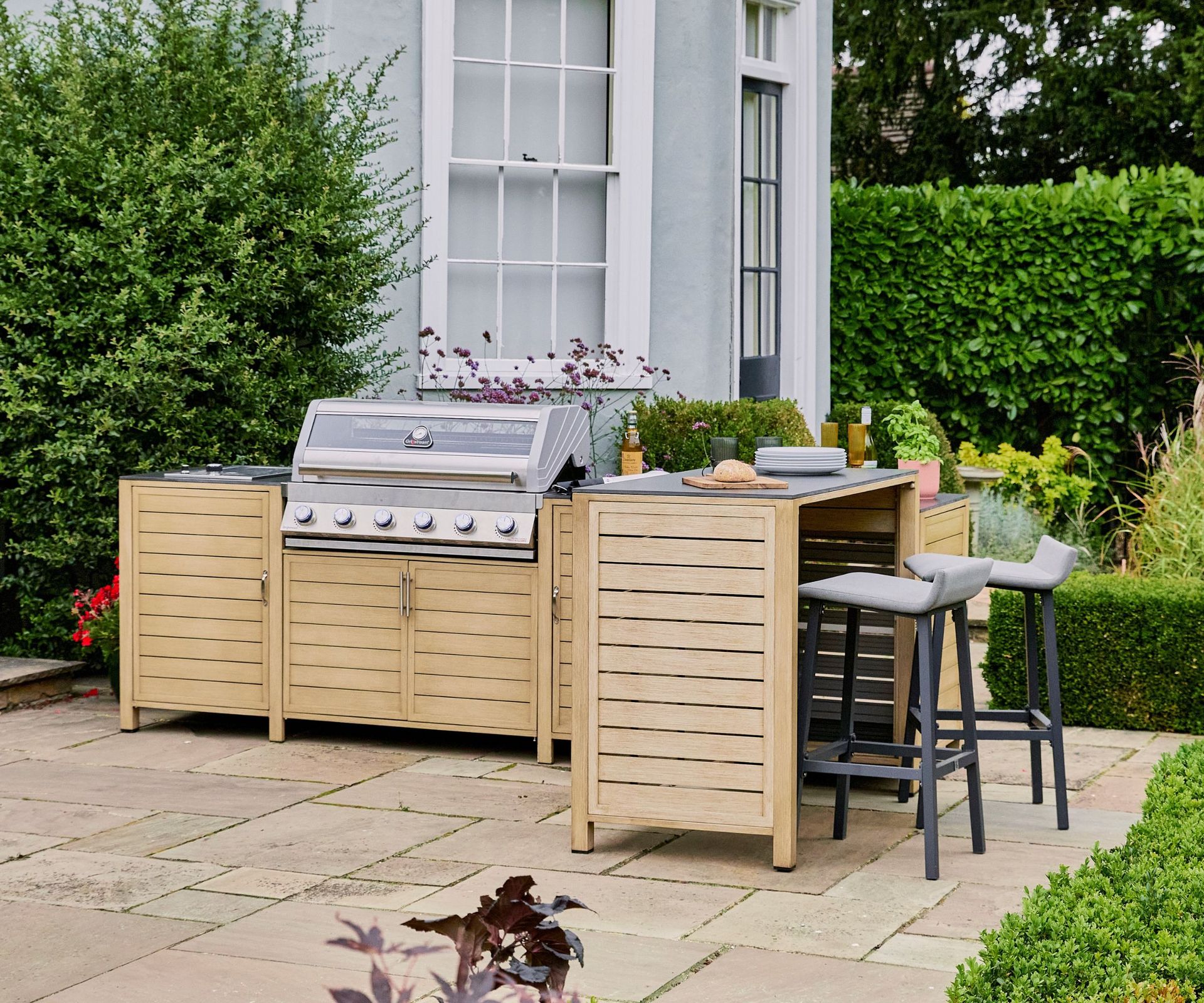 An outdoor kitchen with wooden cabinets, light gray countertops, a stainless steel grill, and a breakfast bar with upholstered stools