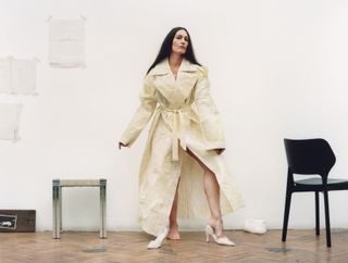 Woman stands in a beige duster coat with beige heels