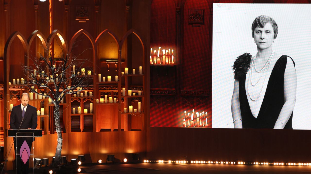 Prince William wearing a dark suit and standing on stage in front of dozens of lit candles and a tree holding cnadles next to a photo of Princess Alice on a large screen