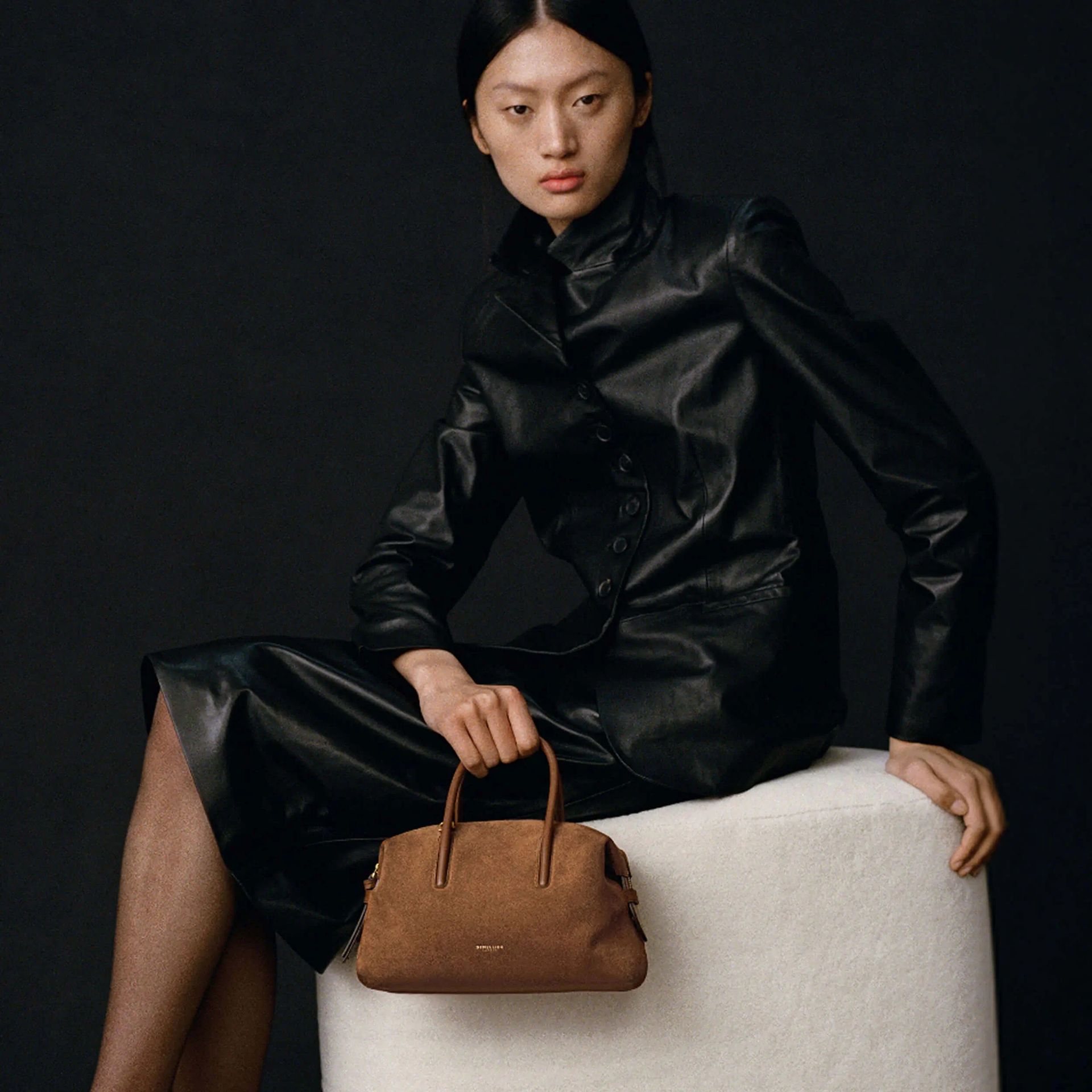 Woman wears black leather jacket and skirt and carries brown suede bag