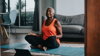 woman in a seated meditative posture on a blue mat in a living room with an ipad device in front of her