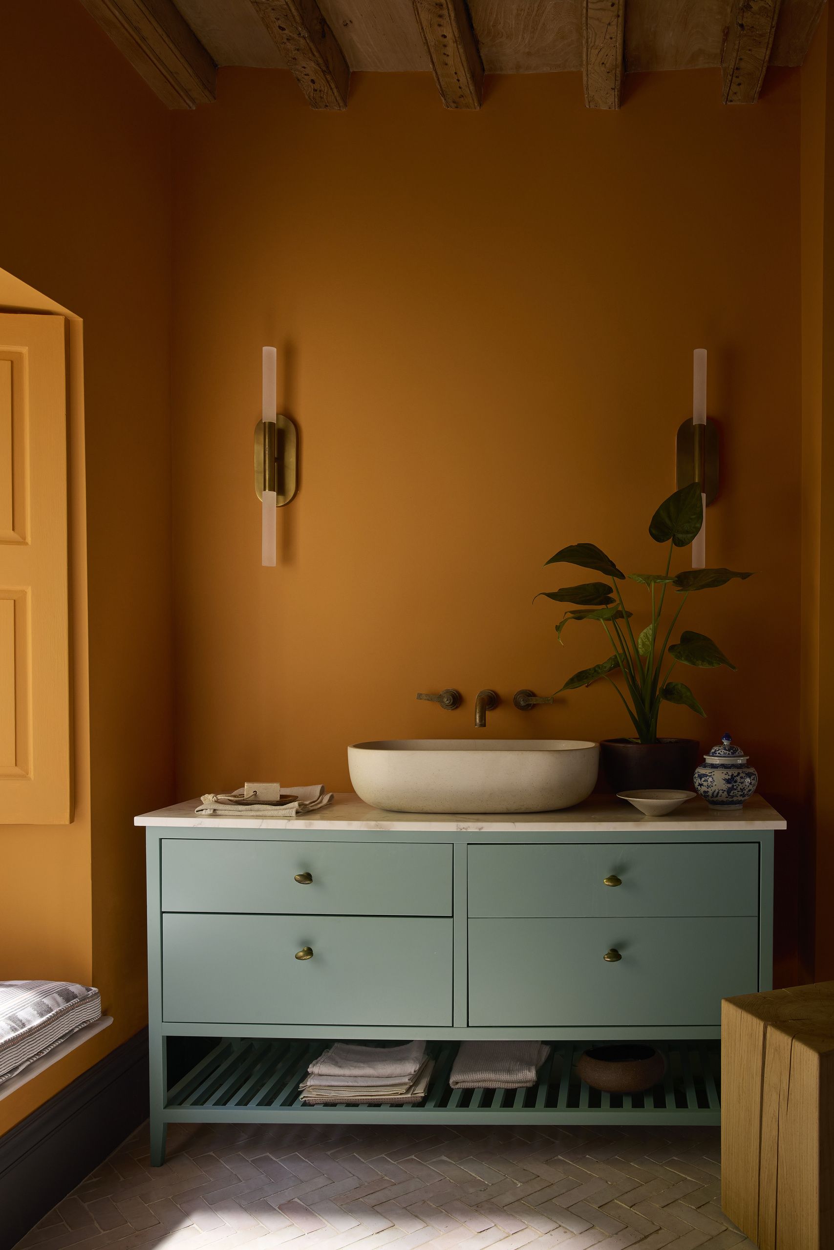 A bright orange bathroom with a light teal vanity with a stone sink and a plant on the counter. There are sconce lights on the wall.