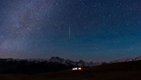 A meteor is pictured streaking vertically Earthwards through a starry night sky. Snow-tipped mountains line the horizon, while illuminated tents are visible below on a grassy plain.