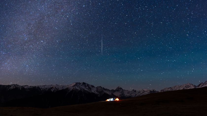 A meteor is pictured streaking vertically Earthwards through a starry night sky. Snow-tipped mountains line the horizon, while illuminated tents are visible below on a grassy plain.