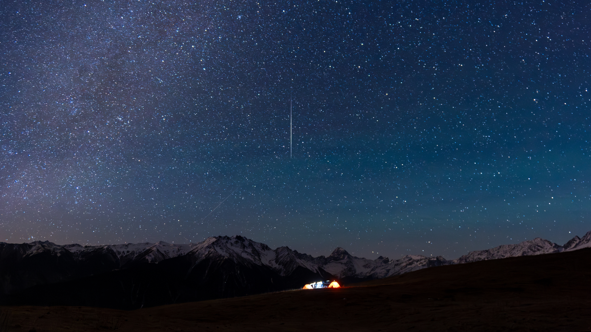 A meteor is pictured streaking vertically Earthwards through a starry night sky. Snow-tipped mountains line the horizon, while illuminated tents are visible below on a grassy plain.