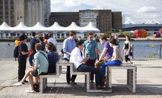 View of a metal and wood panel bench by architecture student Edward Grocott. There are several people sitting on the bench and standing near it. Buildings can be seen in the background along with water at Royal Victoria Docks