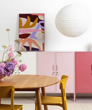 A dining room with white walls with a pink and orange wall art print, a circular pendant light, two pink cabinets one light and one dark, and a wooden circular dining table with yellow chairs and a bouquet