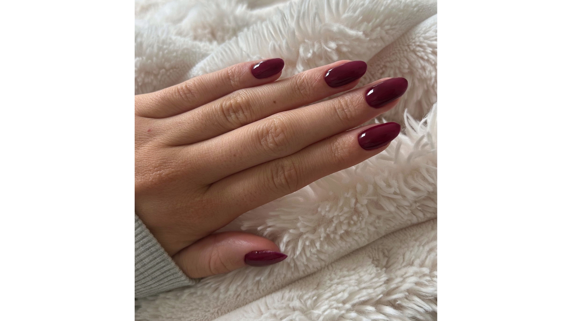 A close-up of Digital Beauty Writer, Sennen Prickett&#039;s hand, which features long, almond-shaped burgundy nails/ pictured above a fluffy cream blanket