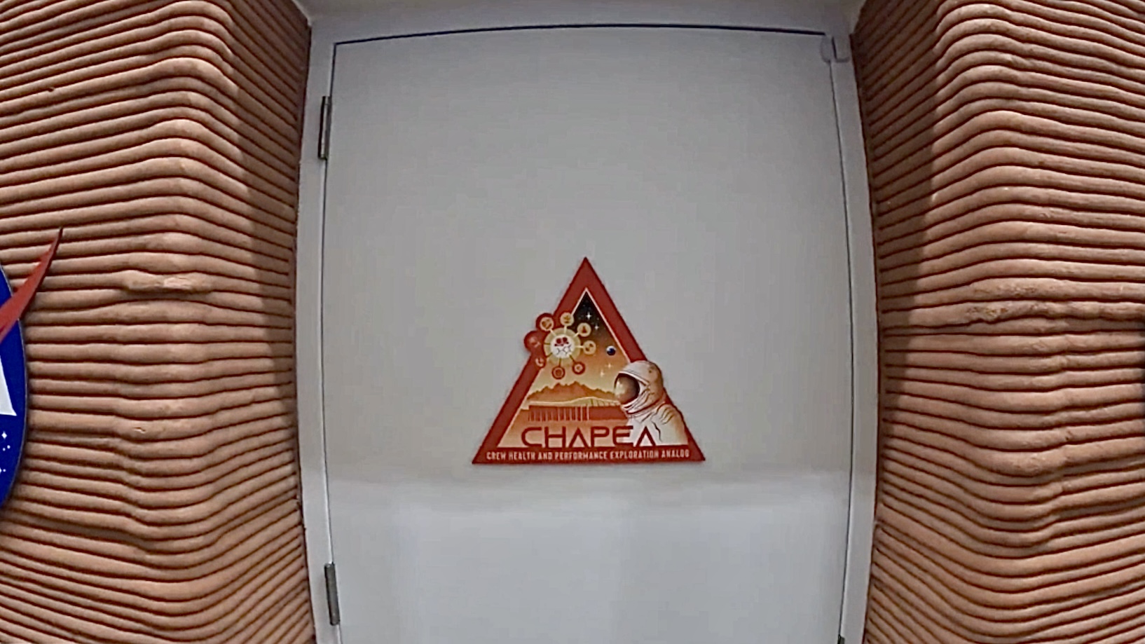 Uma porta branca com o logotipo triangular vermelho da missão CHAPEA no centro.  A entrada e as paredes estruturais são feitas de camadas de argila vermelha impressas em 3D empilhadas.