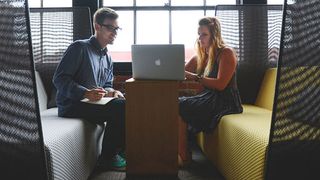 A woman shows a man her laptop screen while he takes notes