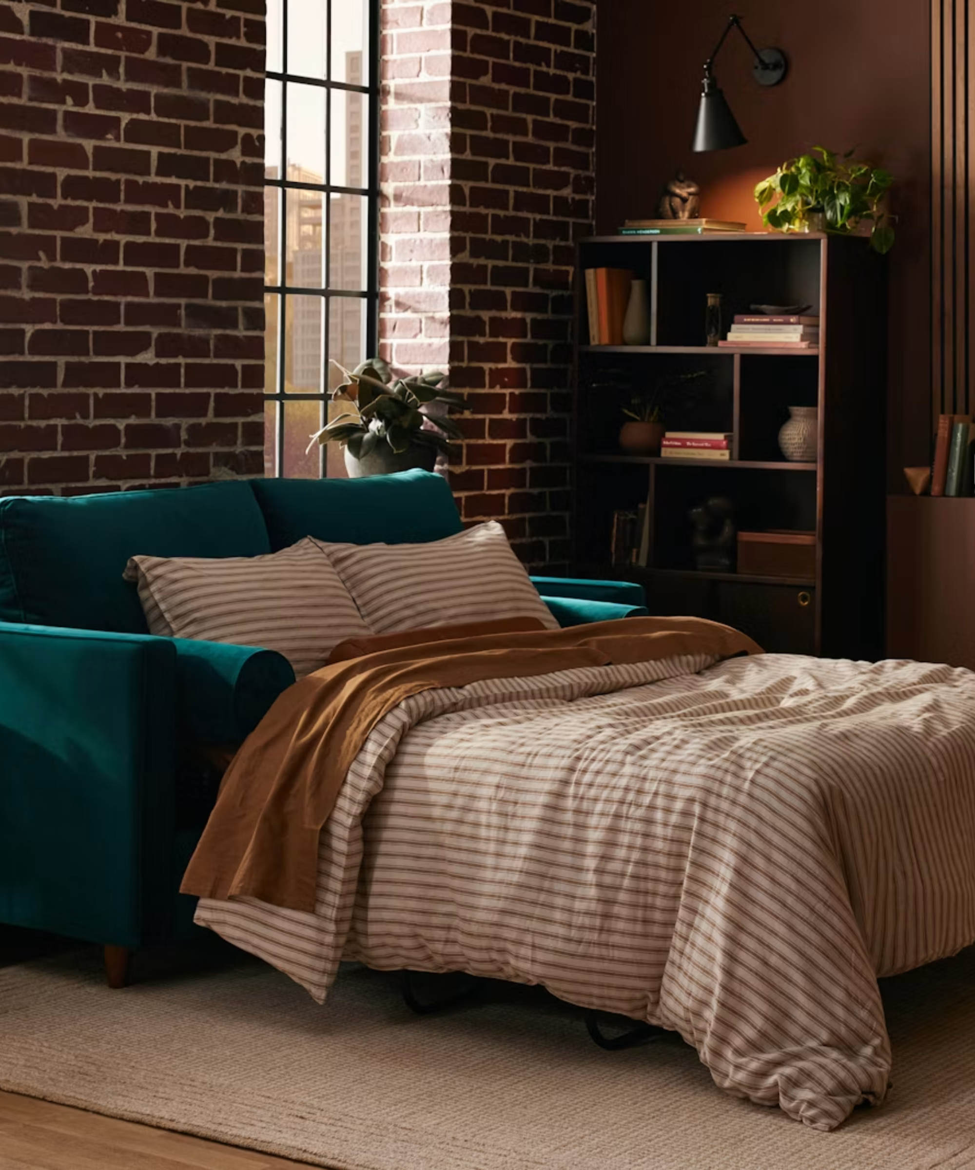 Teal sleeper sofa pictured in a brick-walled urban bedroom with striped bedding and assorted objects