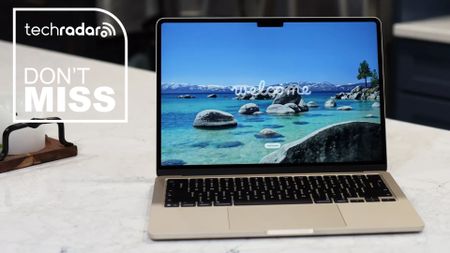 Apple MacBook Air 13-inch (M5) on a table during our review. On the screen is a coastline and the word 'Welcome'. In the upper left corner is a white TechRadar badge that reads 'Don't Miss'