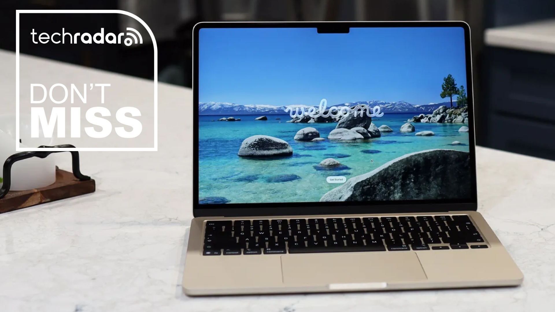 Apple MacBook Air 13-inch (M5) on a table during our review. On the screen is a coastline and the word 'Welcome'. In the upper left corner is a white TechRadar badge that reads 'Don't Miss'