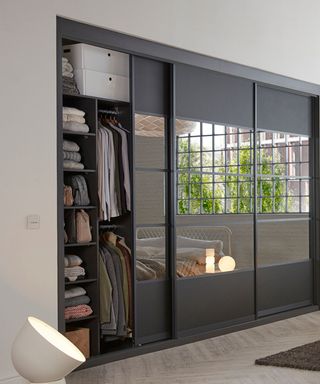 A black wardrobe with three wall storage sliding door panels, with the left hand one open with shelves and rails of shirts hung and folded up, with the doors reflecting a room with a large grid window and a bed