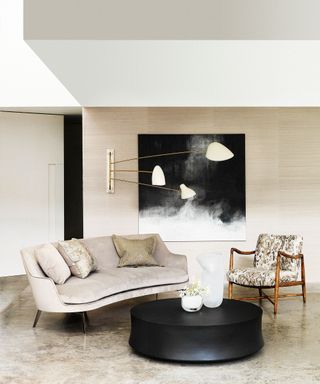 Glamorous neutral living room ideas shown in a beige and gold scheme with curved velvet sofa, black coffee table, modern black and white artwork and statement gold wall lighting.