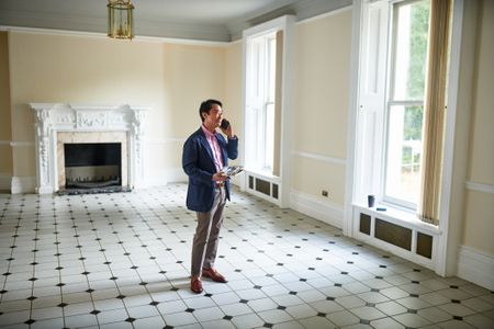 Smartly dressed real estate investor or agent standing in spacious empty room of house for sale talking on cell phone.