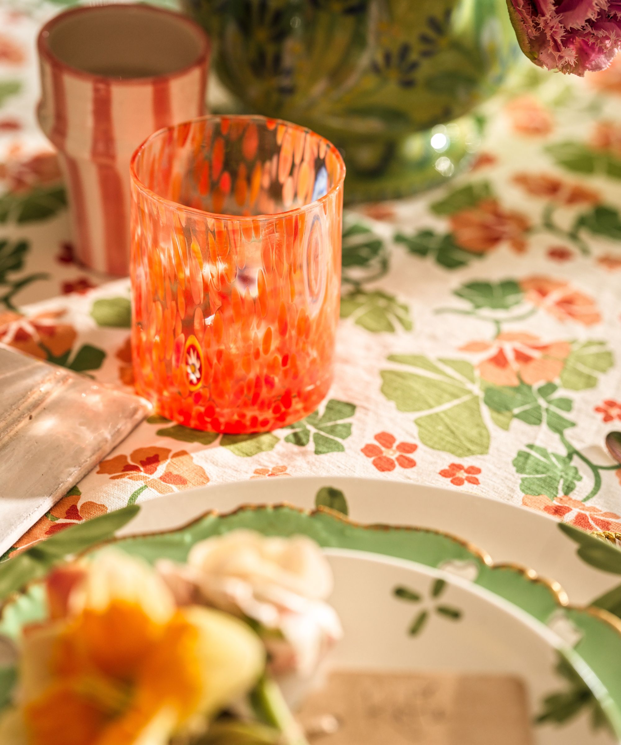 A focused shot of a bright orange speckled glass sitting on a floral tablecloth next to a green-rimmed plate and a striped ceramic tumbler