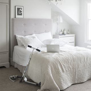 White bedroom, with white headboard, linen, side table and laptop on bed with scooter next to bed
