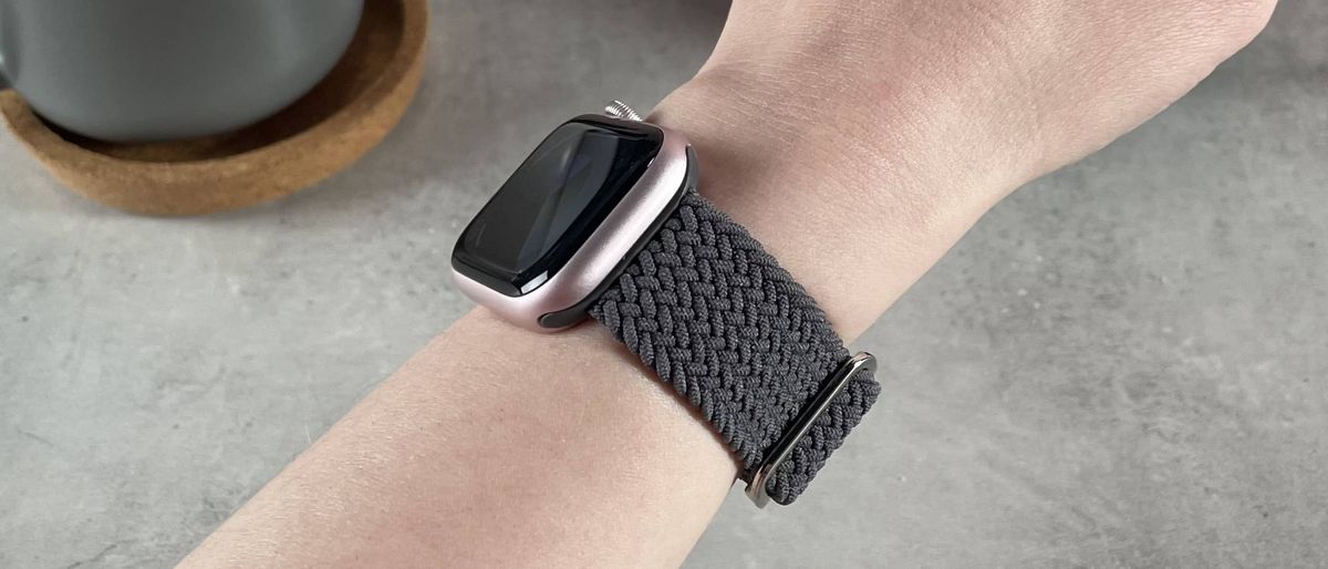 A woman is wearing a pink Apple Watch with a woven grey Apple Watch compatible band.