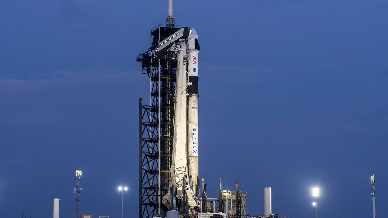 A white and black SpaecX rocket with NASA and SpaceX logos on the side atop a launch pad