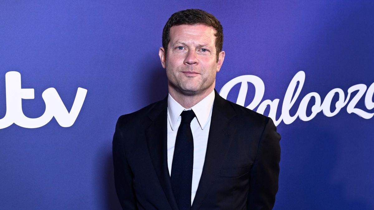 Dermot O&#039;Leary standing in front of a purple background wearing a dark suit and tie