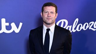 Dermot O'Leary standing in front of a purple background wearing a dark suit and tie