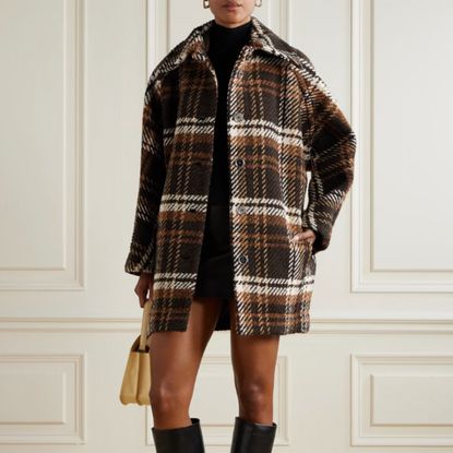 Woman wearing autumn brown and white chequered coat, yellow bag, and black boots, stood in front of white wall. 