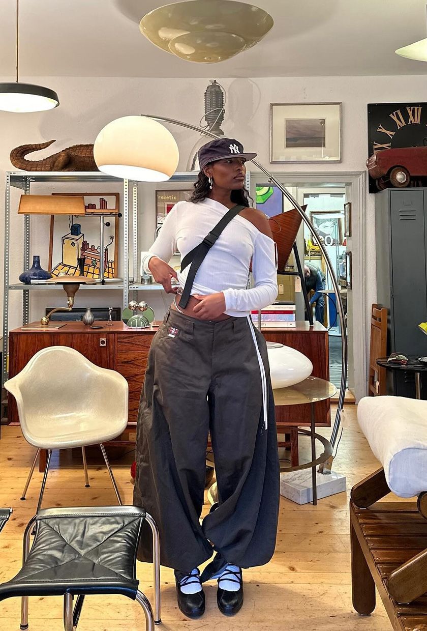 The best ballet sneakers are shown in a photo of a woman standing in a home decor store wearing a navy baseball cap with a white asymmetrical t-shirt and gray baggy cargo pants, white socks, and black lace-up ballerina sneakers