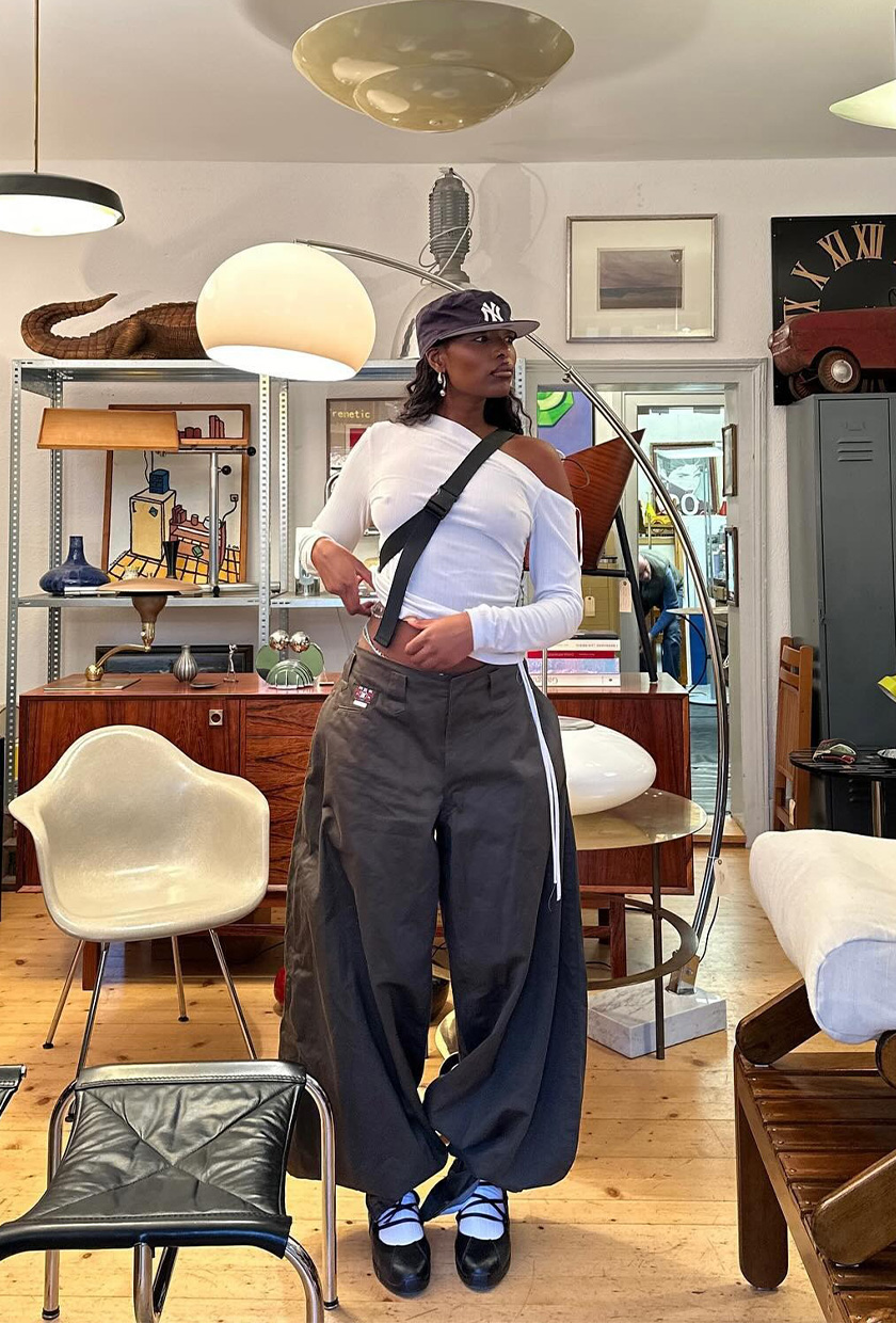 The best ballet sneakers are shown in a photo of a woman standing in a home decor store wearing a navy baseball cap with a white asymmetrical t-shirt and gray baggy cargo pants, white socks, and black lace-up ballerina sneakers
