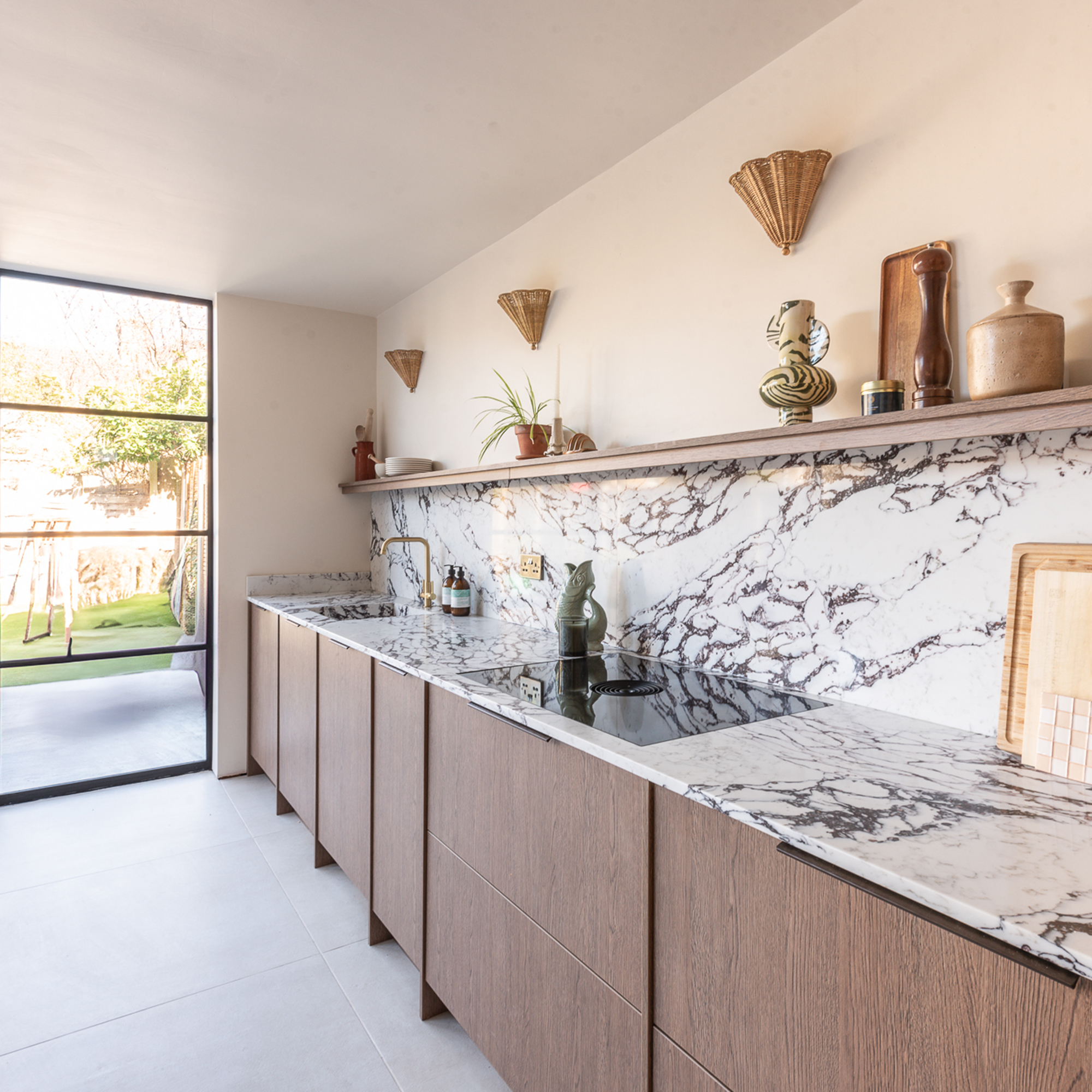Oak kitchen with black and white marble worktop and splashback up to an open shelf with wicker wall lights on the wall above